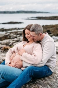 Expectant couple sitting on rocks at Newcastle Commons beach during their maternity session.